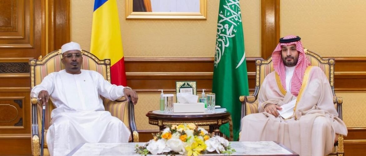 Two men in traditional white robes sit in gold-trimmed chairs at a formal meeting, with a floral centerpiece and national flags behind them.