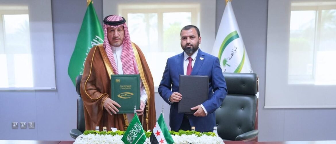 Two men in formal dress stand beside a table with documents during a signing ceremony, with Saudi and other flags in the background.