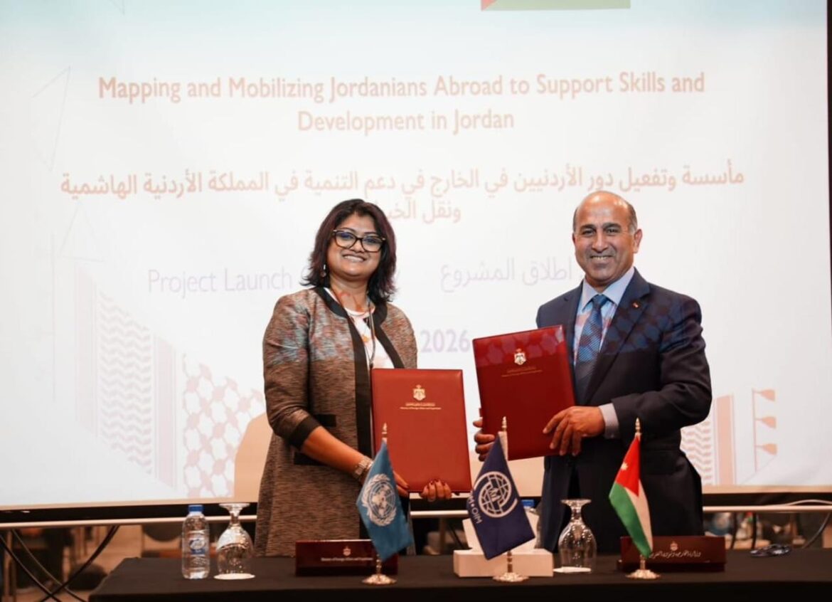Two presenters hold red folders at a formal signing ceremony, with UN and Jordan flags on the table and a projected project title behind them.