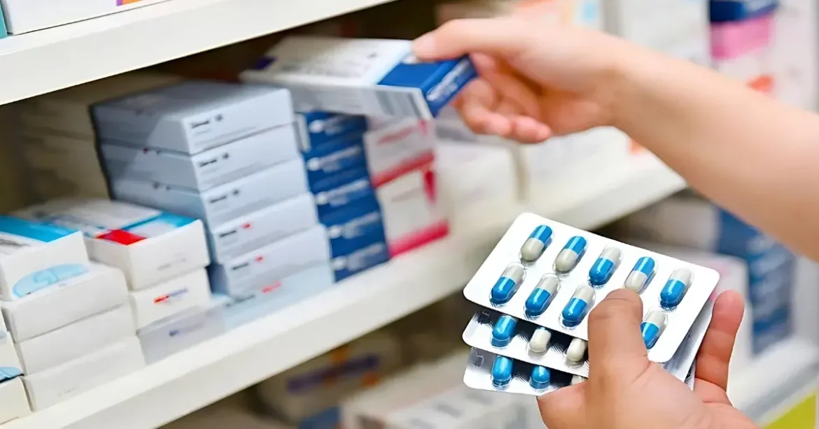 Person selecting prescription drugs from a pharmacy shelf, holding blister packs of blue-and-white capsules in the foreground