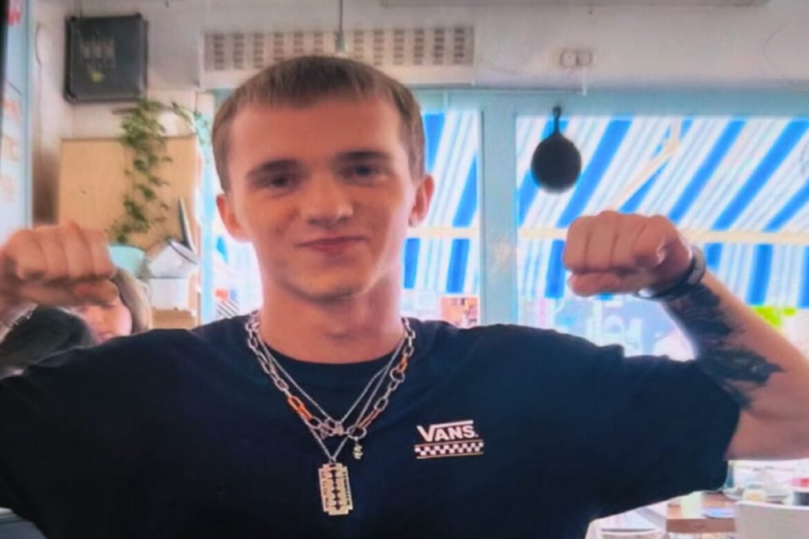 Man in a black Vans shirt with multiple chains raises both fists in a celebratory pose indoors, blue striped windows in the background.