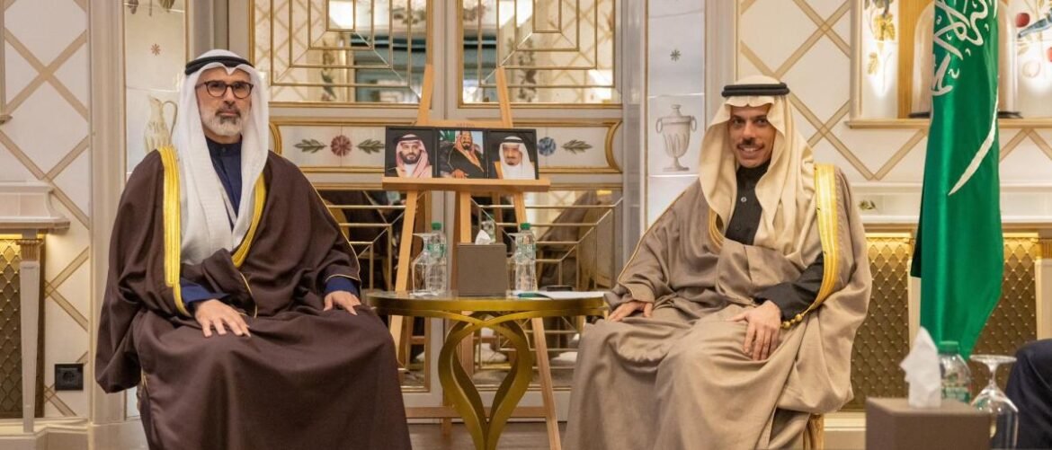 Two men in traditional Saudi thobes and ghutras sit on ornate chairs in a decorated room, with a small table between them and a Saudi flag nearby.