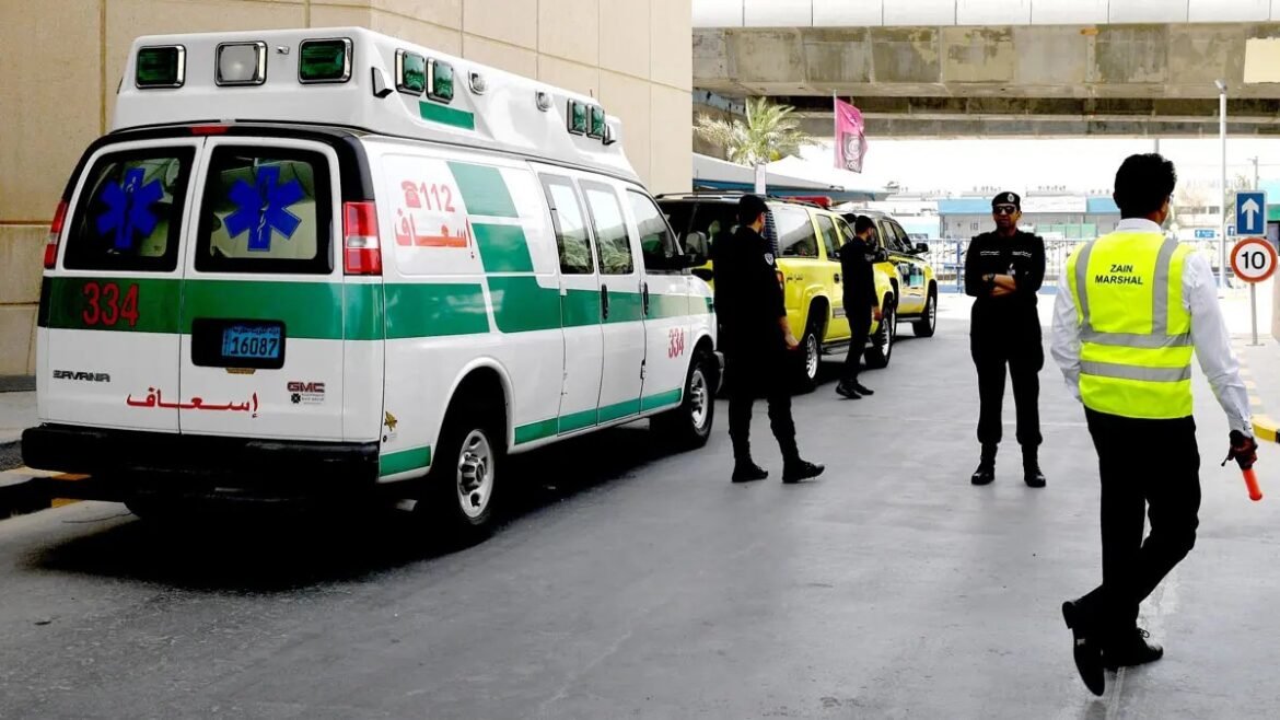 White ambulance with green stripe and Arabic text parked at a curb, with uniformed staff nearby and a marshal in a high‑visibility vest nearby outside a building.
