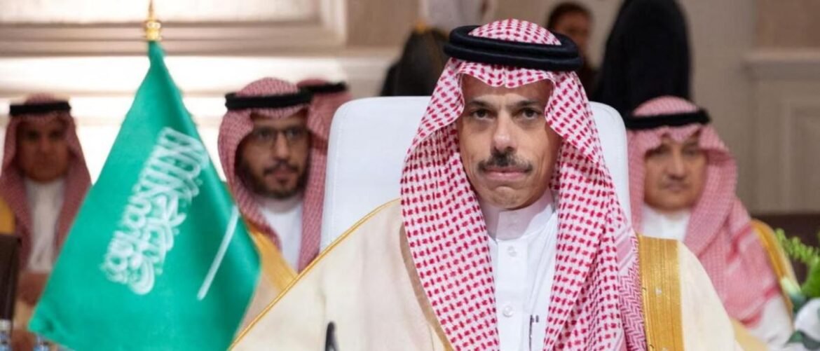 Man in traditional Saudi dress seated at a formal event, with a green Saudi flag nearby and other attendees in the background.