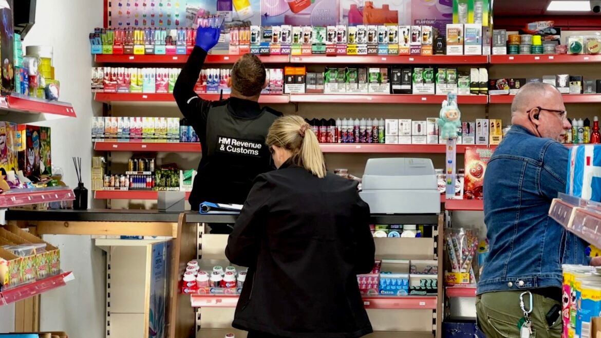 Cashier area in a colorful shop with two staffers behind the counter; one wearing an HM Revenue & Customs jacket reaches up to a top shelf while a customer stands nearby.