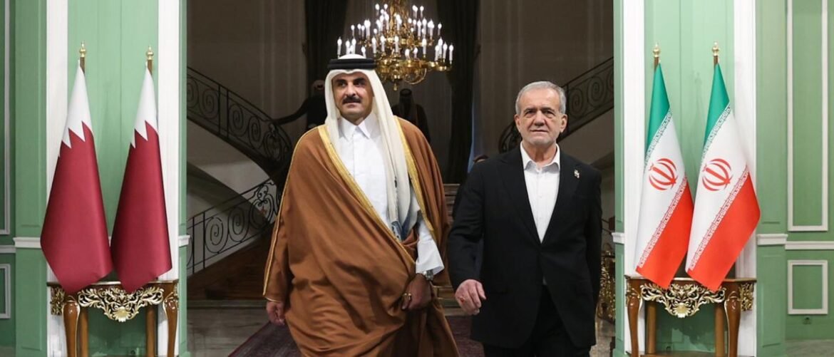 Two men in formal attire walk side by side through a grand green hallway, flanked by Qatari and Iranian flags.