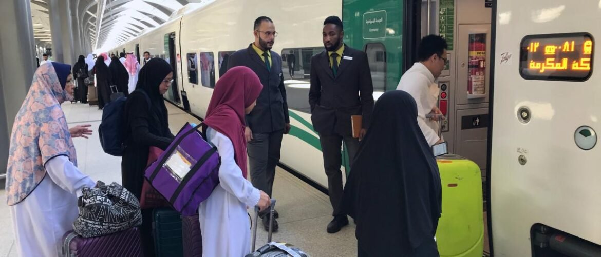Travelers with rolling suitcases line up at a bright platform to board a white train with green stripes, while two uniformed staff members oversee the boarding.