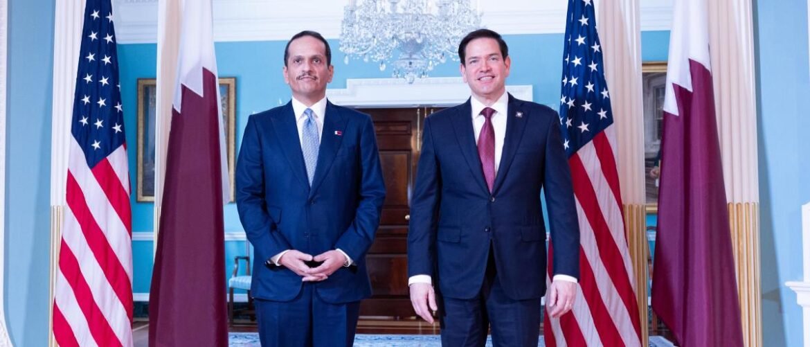 Two men in dark suits stand side by side in a formal doorway, flanked by U.S. and national flags in a bright blue room, posing for a diplomatic photo.
