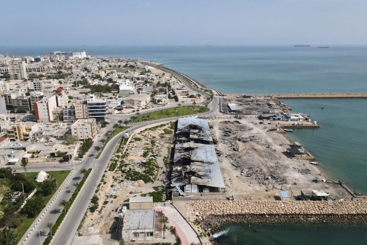 Aerial view of a coastal city with a curved breakwater, mid-rise buildings, and damaged waterfront structures along the beach.