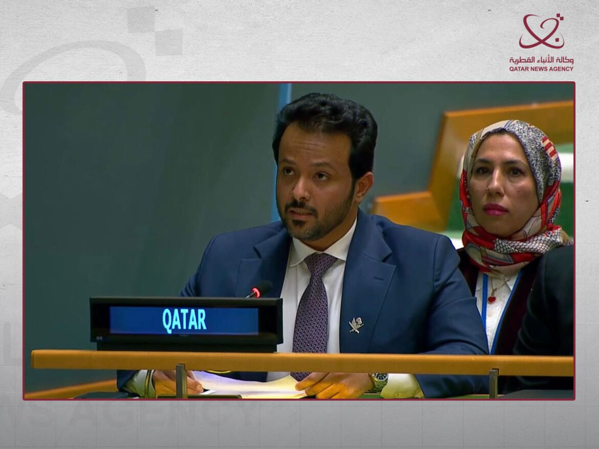 Man in a blue suit speaks into a microphone at a podium labeled 'QATAR', with a woman in a patterned hijab seated beside him in a formal meeting.