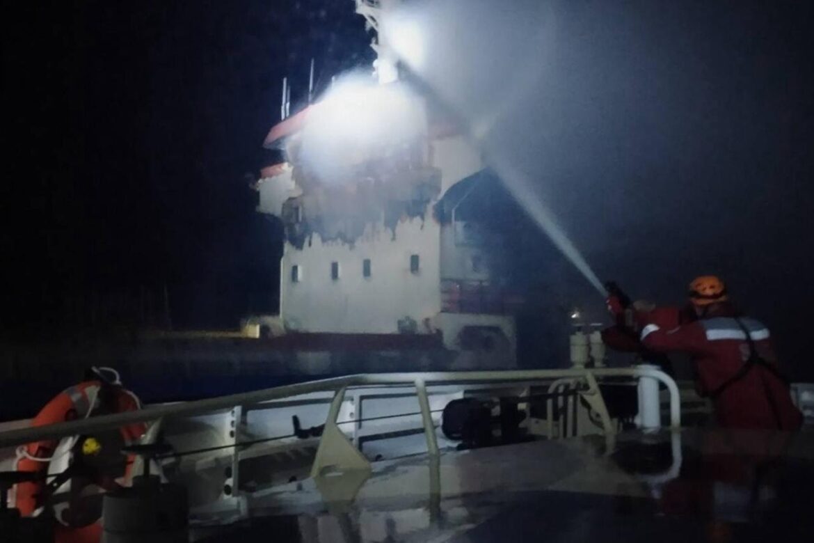 Nighttime on a ship deck as firefighters in red suits spray a powerful stream of water at a white, damaged building.