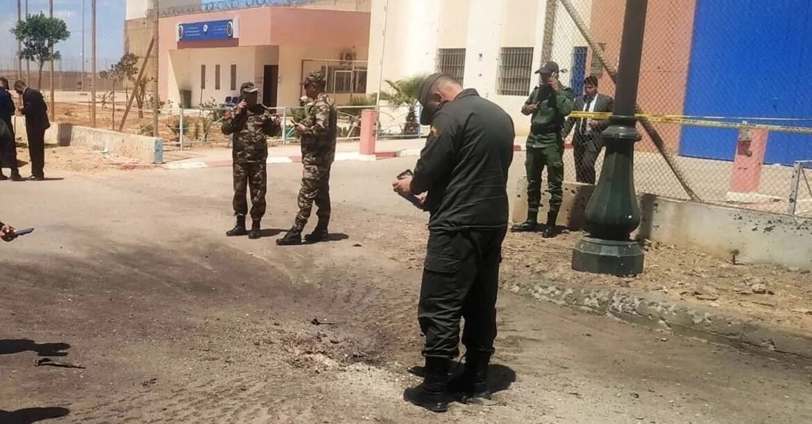 Security personnel in camouflage and black tactical gear inspect a damaged road outside a fenced building, with yellow tape in the background.