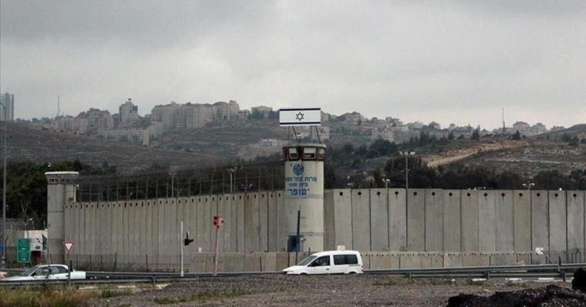 Concrete border fence with a guard tower flying an Israeli flag; a white van on the highway in front.
