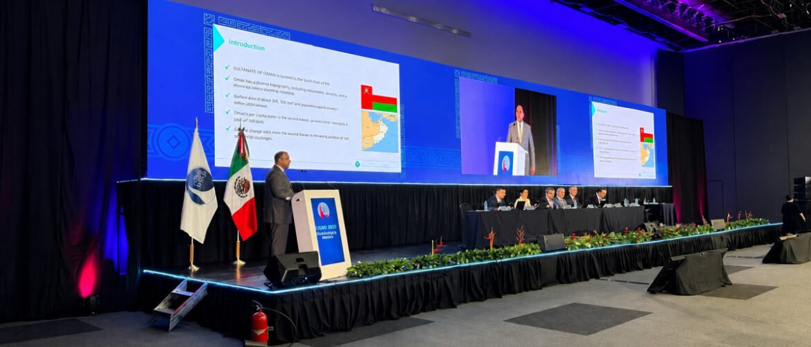 Speaker at a podium presenting to a panel on a long stage, with large screens displaying slides and flags behind the speaker.