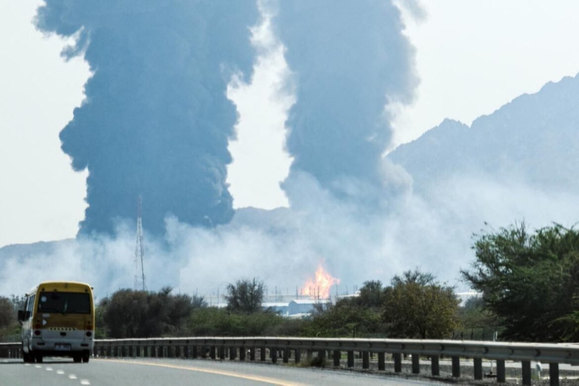 Yellow bus on a highway as large dark smoke plumes rise from a distant fire near the hills, flames visible on the horizon.