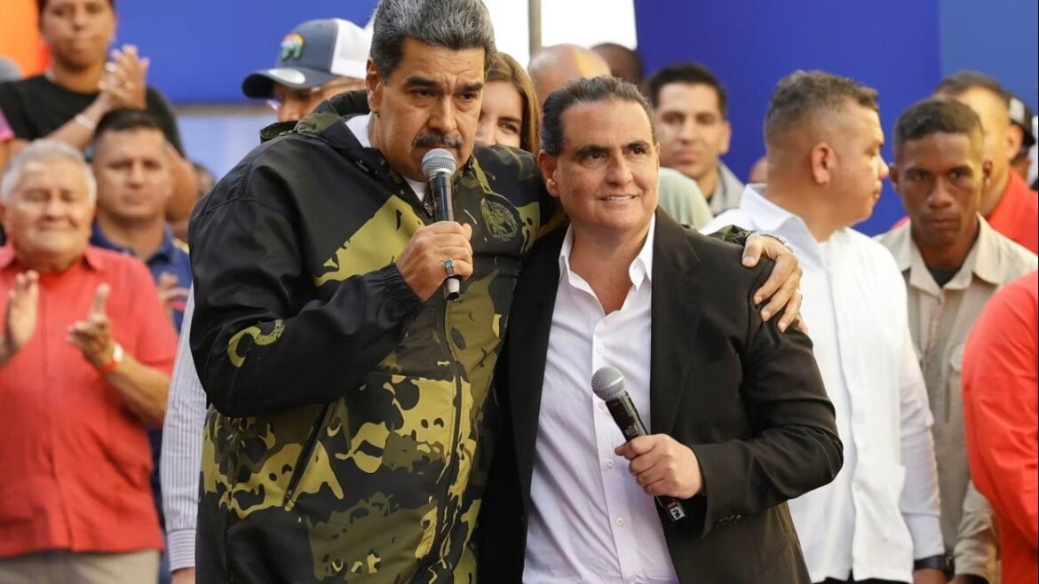 Two men on a stage with microphones, one in a camouflage jacket and one in a suit, smiling with a cheering crowd behind them.