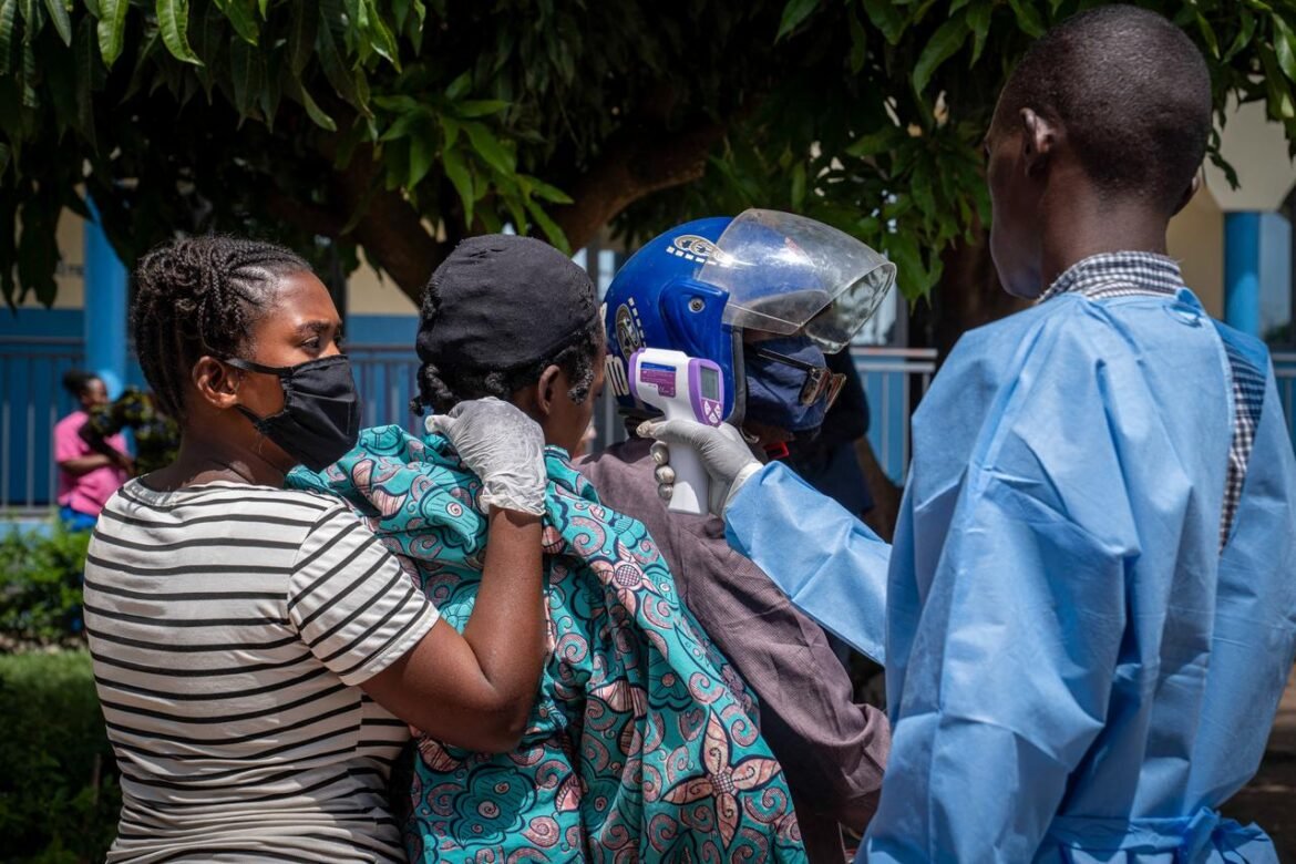 Healthcare worker in blue protective gown uses a handheld thermometer to take a woman's temperature outdoors; all wear masks and gloves nearby.