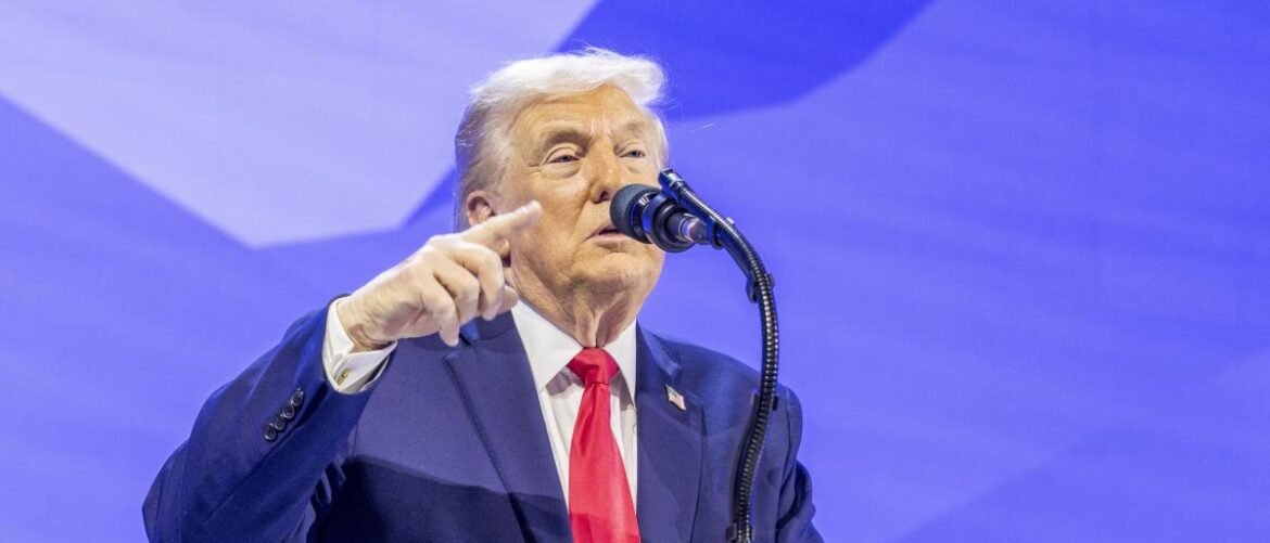 Man in a navy suit and red tie speaks into a microphone on stage, gesturing with his finger on a blue backdrop field.