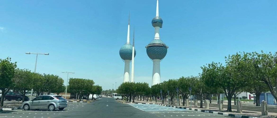 Kuwait Towers rising above a tree-lined street on a sunny day with parked cars in the foreground.