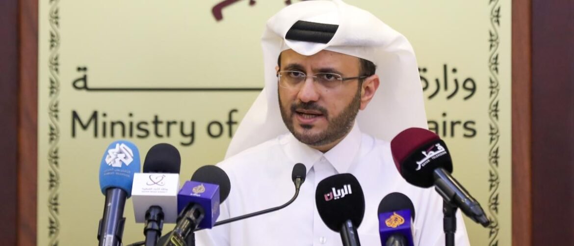 Qatari official wearing a white thobe and ghutra speaks at a ministry press conference, with several media microphones in front.