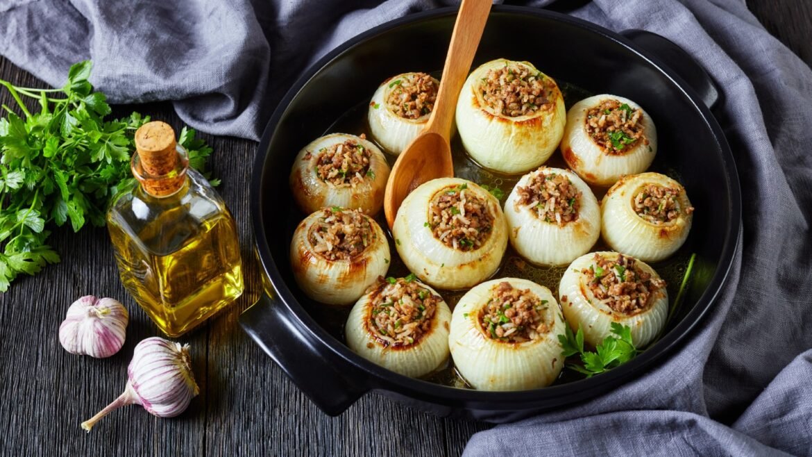 Skillet of roasted onions stuffed with seasoned ground meat, garnished with herbs, with a bottle of olive oil and garlic on a dark wooden table.