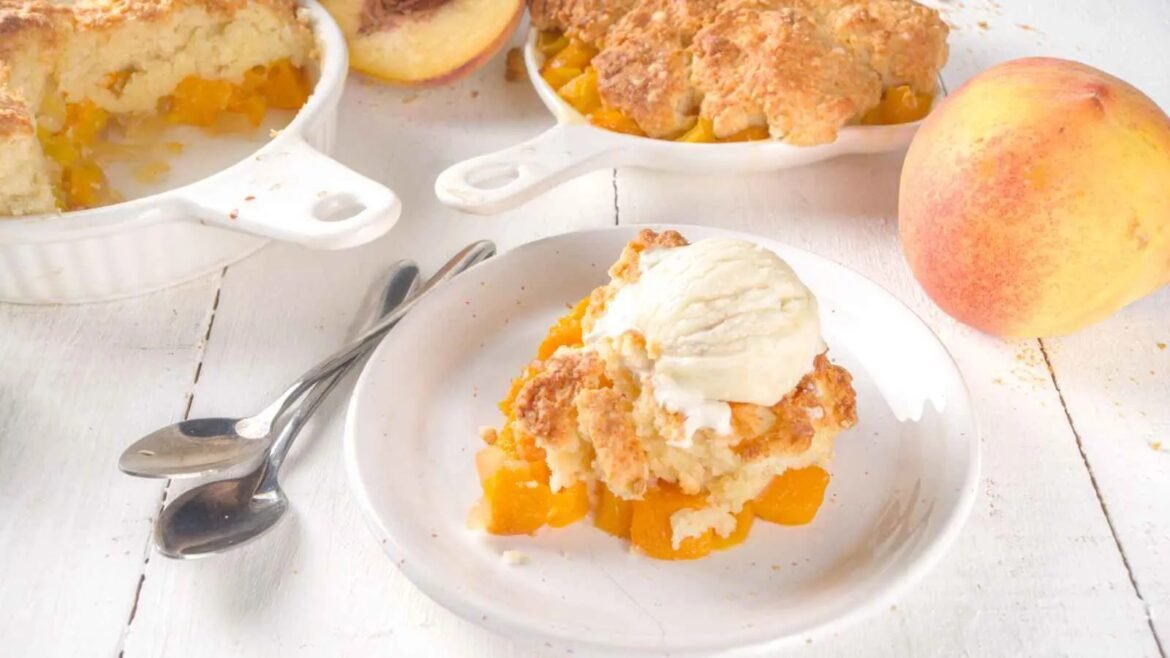 Peach cobbler with vanilla ice cream on a white plate, peach chunks visible and a white baking dish in the background on a white wooden table