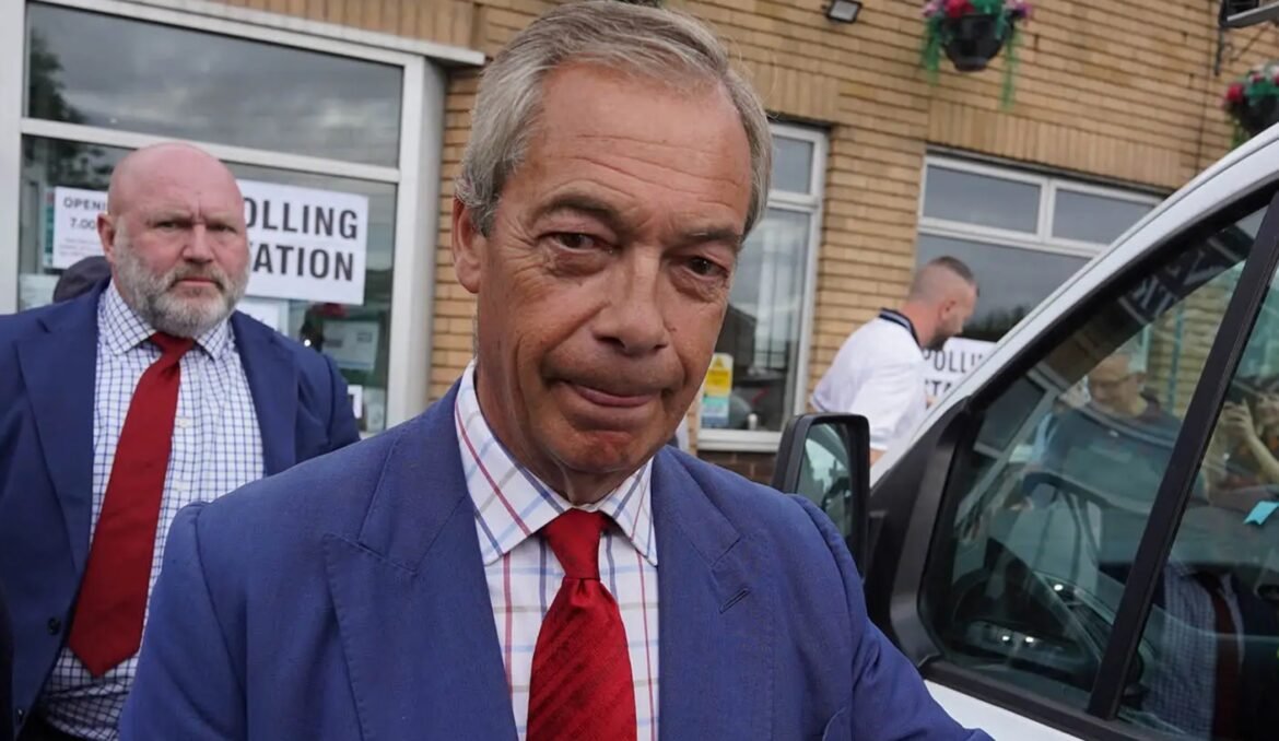 Man in a blue suit and red tie standing outside a polling station, with a ‘Polling Station’ sign visible in the window behind him.