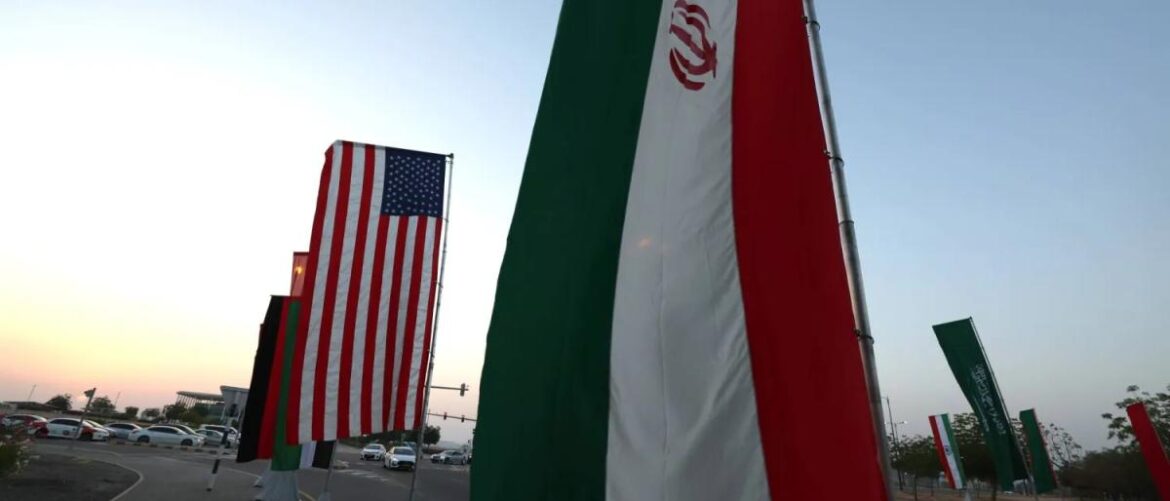 Several tall national flags flutter in a parking lot at sunset, including the United States and Iran flag(s).