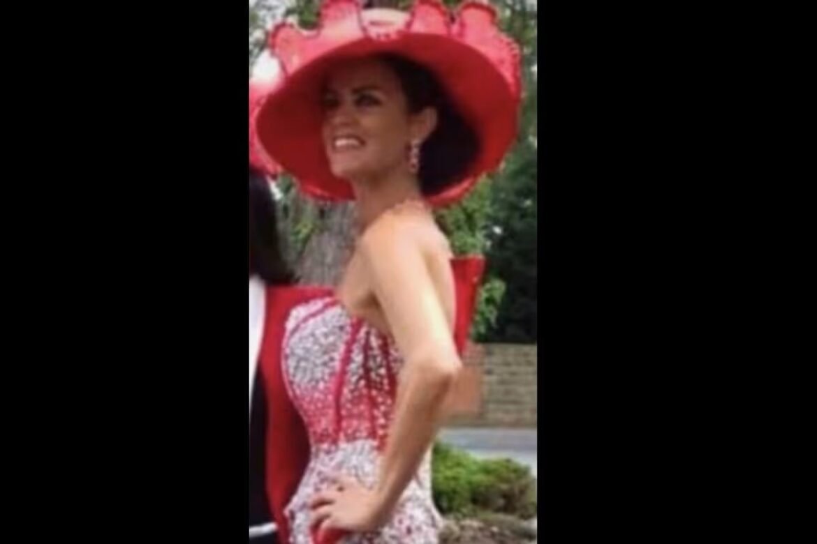 Woman in a large red hat and red-and-white strapless dress posing outdoors with hand on hip, smiling slightly.