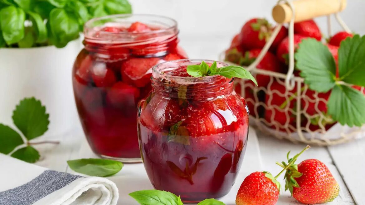 Two glass jars of strawberry jam with fresh basil garnish on a white surface, surrounded by strawberries and a basket of berries in the background.