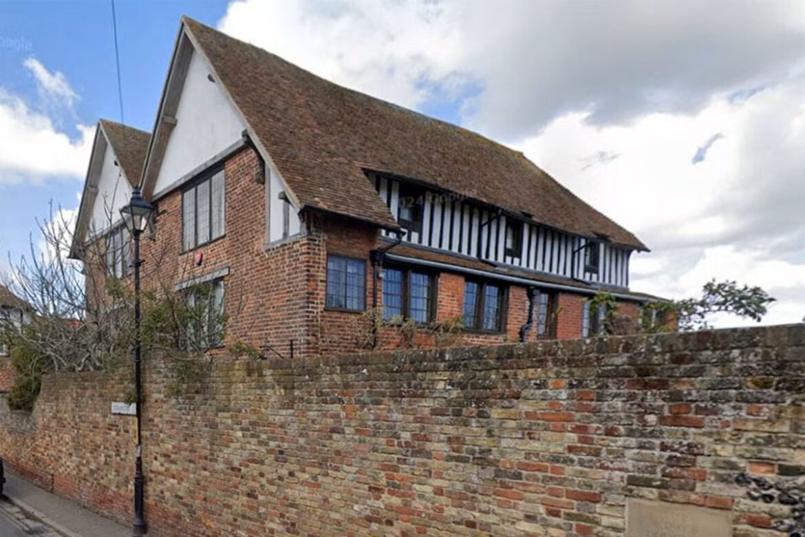Brick building with a timber-framed upper facade behind a low brick wall on a sunny street, with a vintage lamppost nearby.