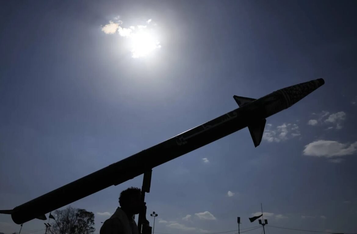 Silhouetted person carrying a large rocket on a launcher against a bright sunlit sky and scattered clouds in the background.