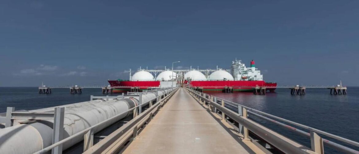 Concrete pier with metal railings leads to a large red-and-white LNG carrier with white spherical tanks at the end of calm water.