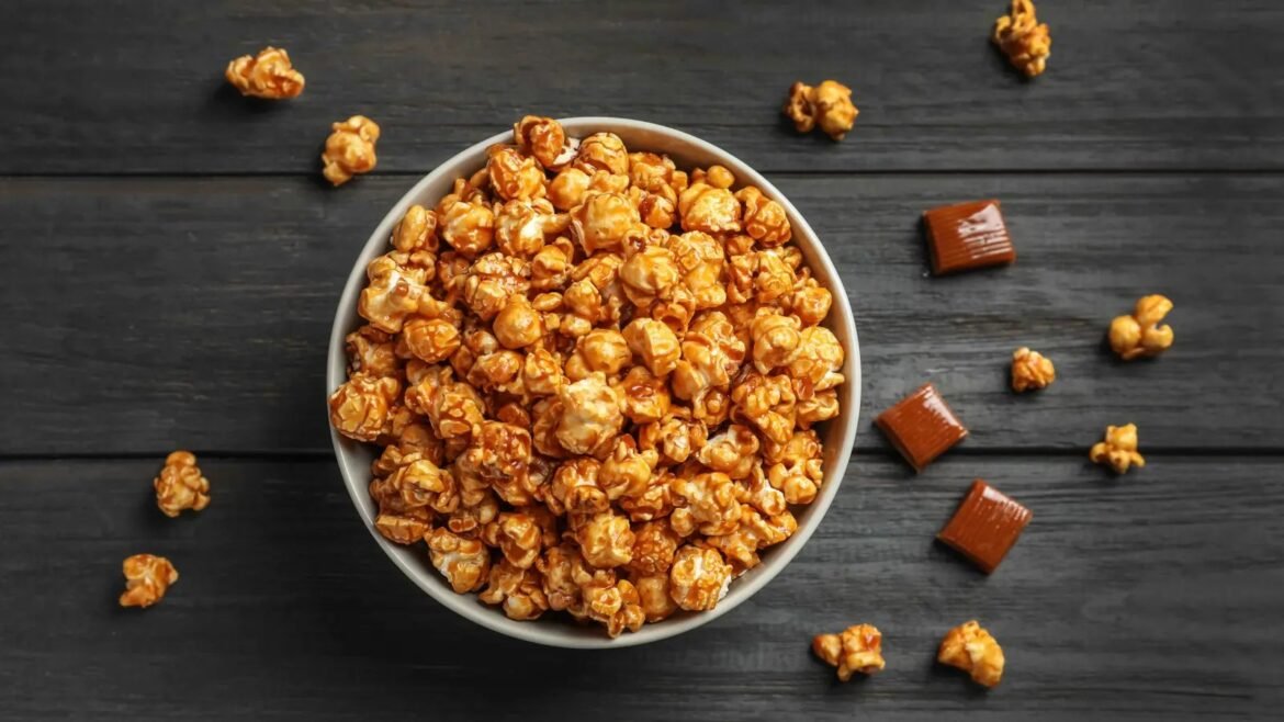 Bowl of caramel popcorn on a dark wooden table with a few scattered popcorn pieces and chocolate squares nearby