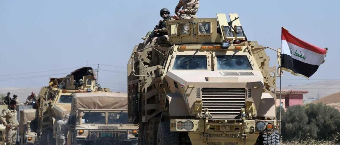 Armored military convoy driving through a desert, soldiers on top of the lead vehicle with an Iraqi flag flying from the front.