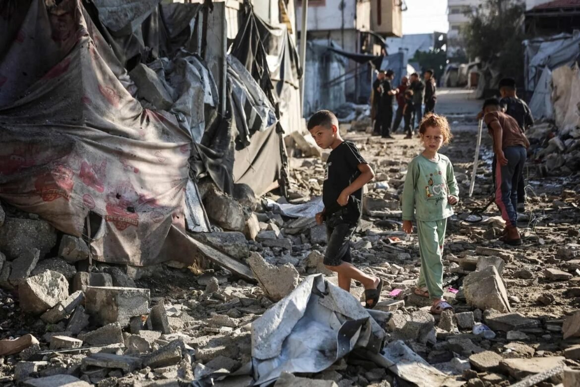 Children stand among rubble in a destroyed street, tents visible in the background, conveying life amid conflict.