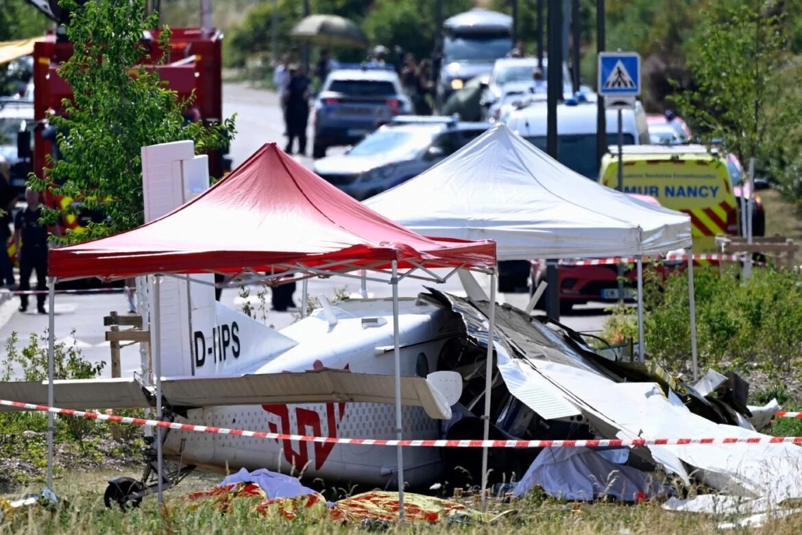 Small crashed airplane under red and white tents at a cordoned incident scene, emergency responders in the background.