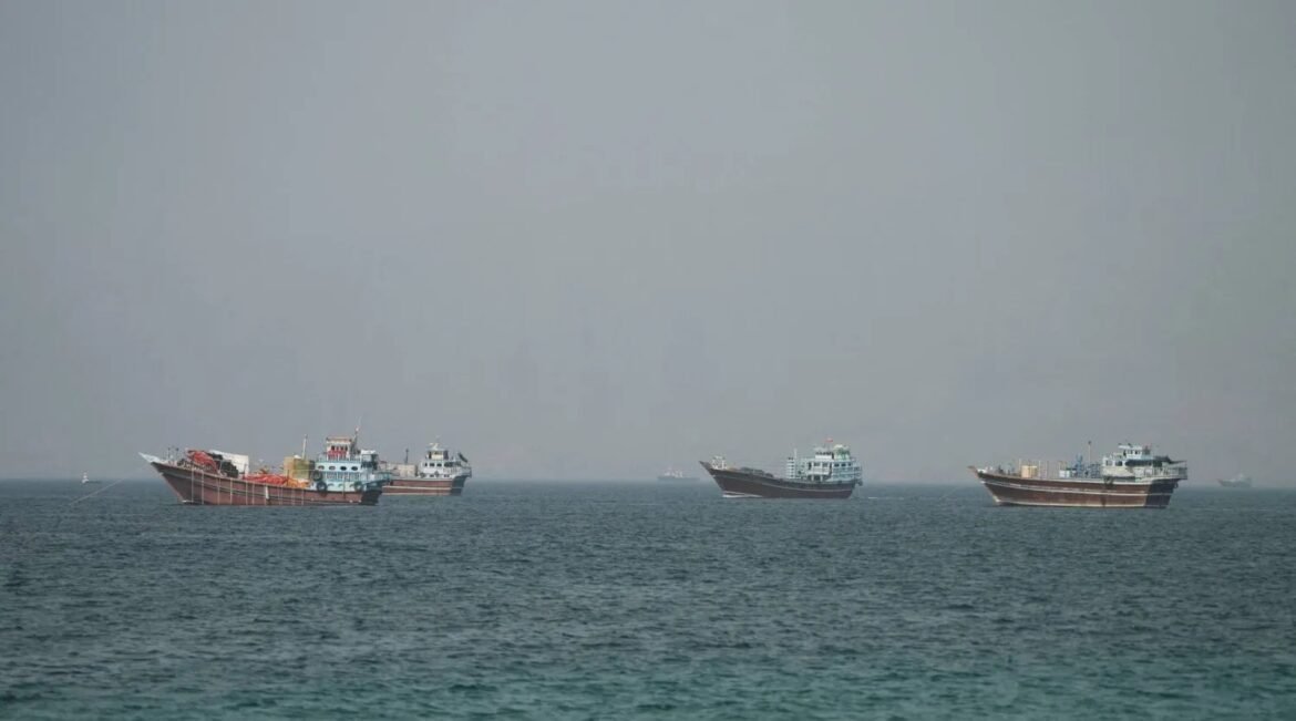 Several large cargo ships floating on a calm sea under a hazy sky, spaced along the horizon far from shore, each vessel loaded with containers and equipment.