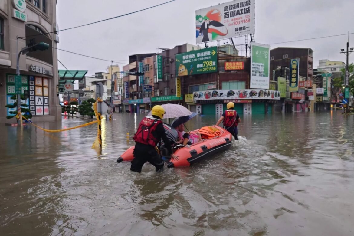 Rescue team wades through a flooded city street guiding an orange inflatable boat with passengers and a person holding an umbrella. Empty storefronts line the waterlogged avenue beneath cloudy skies.