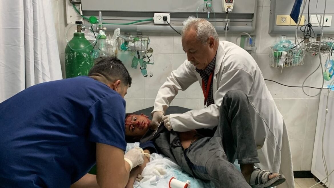 Two clinicians treat a man with facial injuries on a hospital bed in a medical room with equipment around them.