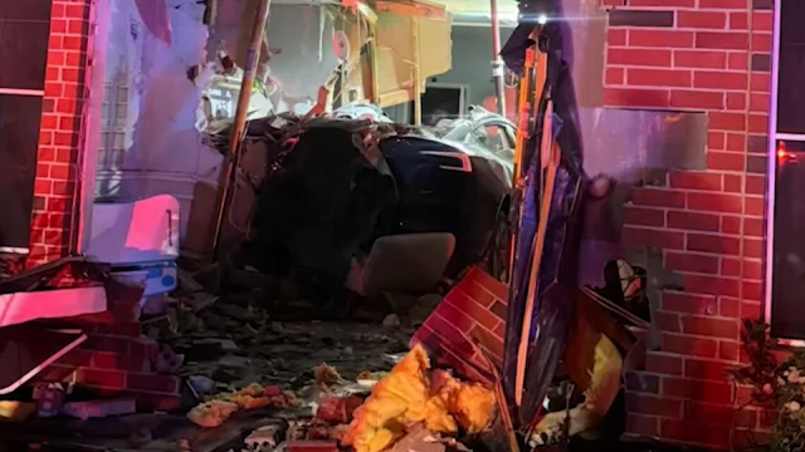 Car crashed into a brick storefront, debris and insulation scattered on the ground, emergency lights glow red.
