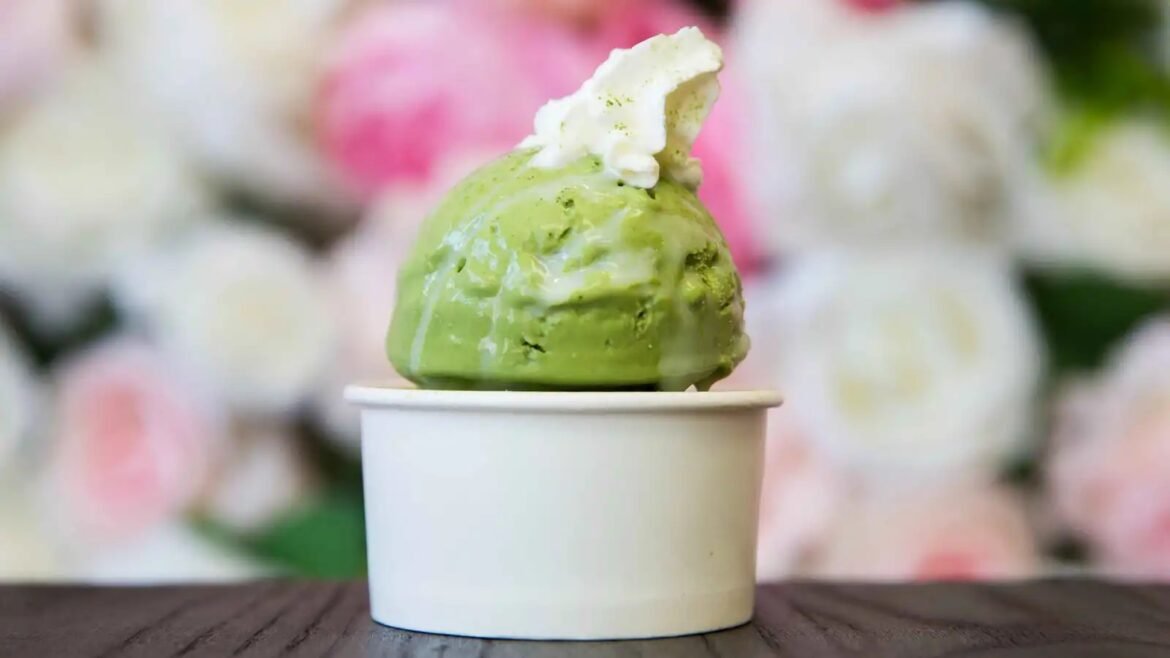 Scooped green ice cream in a white cup with a small white wafer on top, blurred floral background.