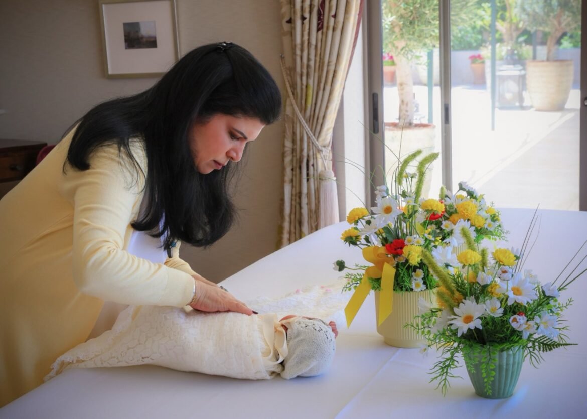 Woman smoothing white lace fabric on a table, with a yellow-and-white flower arrangement nearby by a window