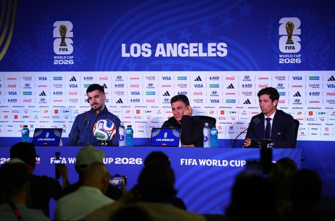 Panel of three men at a FIFA World Cup 2026 press conference in Los Angeles, with microphones and a football on the table, audience in the foreground and a blue backdrop full of sponsor logos.