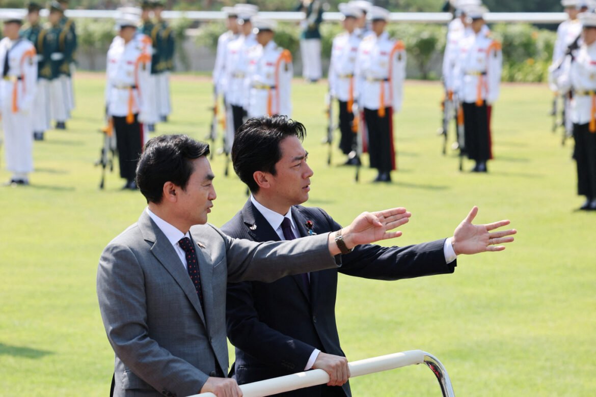 Two men in suits stand at a railing, one gesturing with his arm extended while a line of uniformed guards stands in formation on a grassy field behind them.