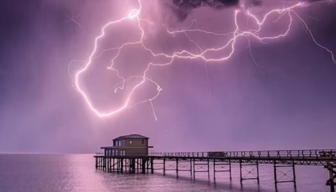 Lightning bolts streak across a purple stormy sky over a wooden pier extending into calm water, ending with a small building at the end.