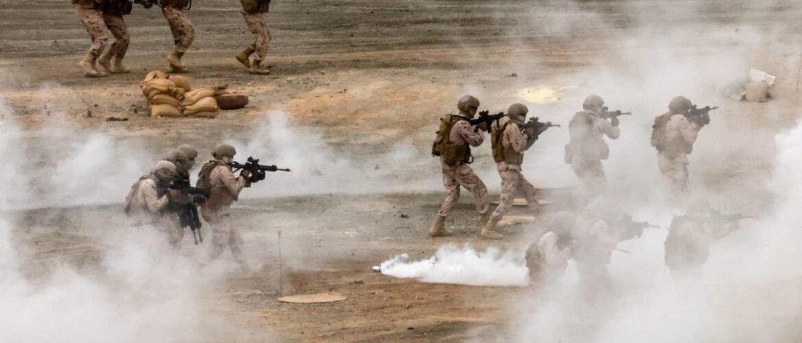 Group of soldiers in desert gear advancing through smoke with rifles during a training exercise, on a dusty range.