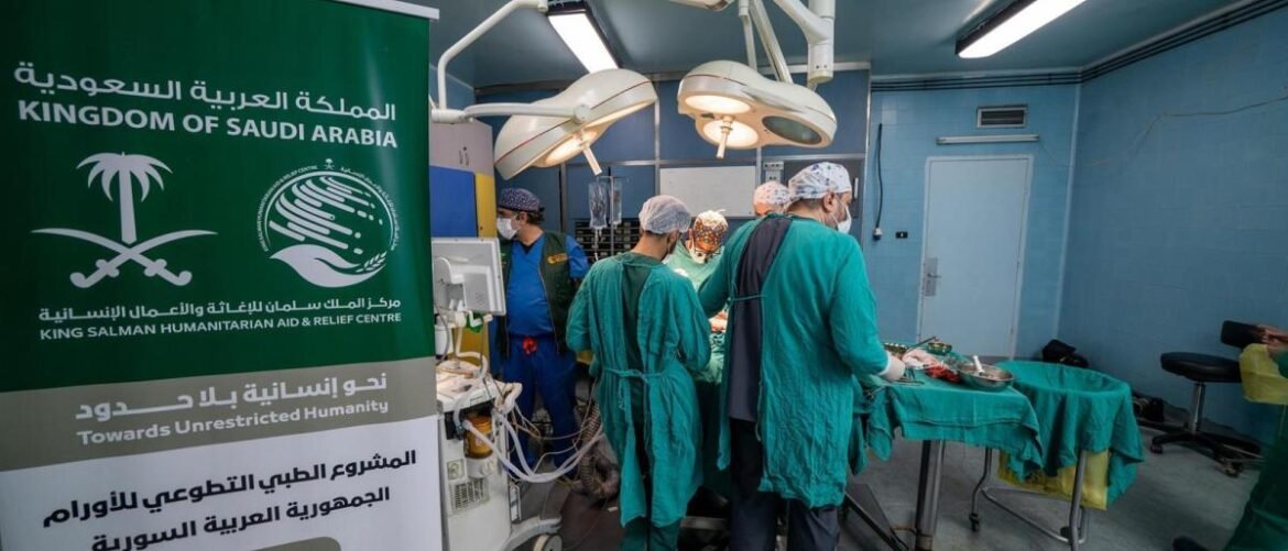 Operating room with surgeons in green scrubs performing surgery; a large banner reads Kingdom of Saudi Arabia and King Salman Humanitarian Aid & Relief Centre in the foreground.