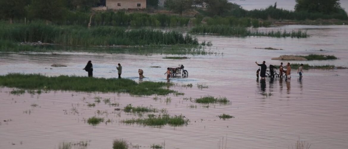 Group of people wading through knee-deep floodwater near a rural area; a bicycle is partly submerged.