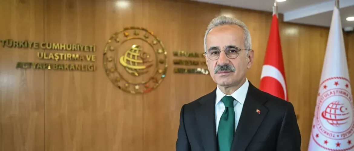 Formal portrait of a man in a dark suit and green tie, standing in front of a wooden wall with an official seal and Turkish flag on his right.